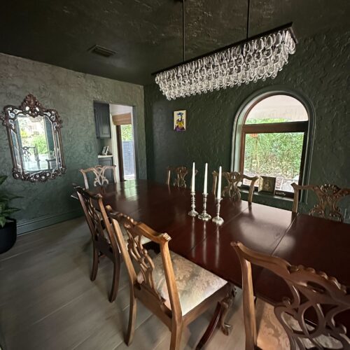 A dark green dining room with a long wooden table, eight ornate chairs, a crystal chandelier, a mirror on the wall, three candles on the table, and a large arched window with greenery outside.