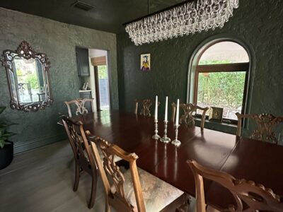 A dark green dining room with a long wooden table, eight ornate chairs, a crystal chandelier, a mirror on the wall, three candles on the table, and a large arched window with greenery outside.