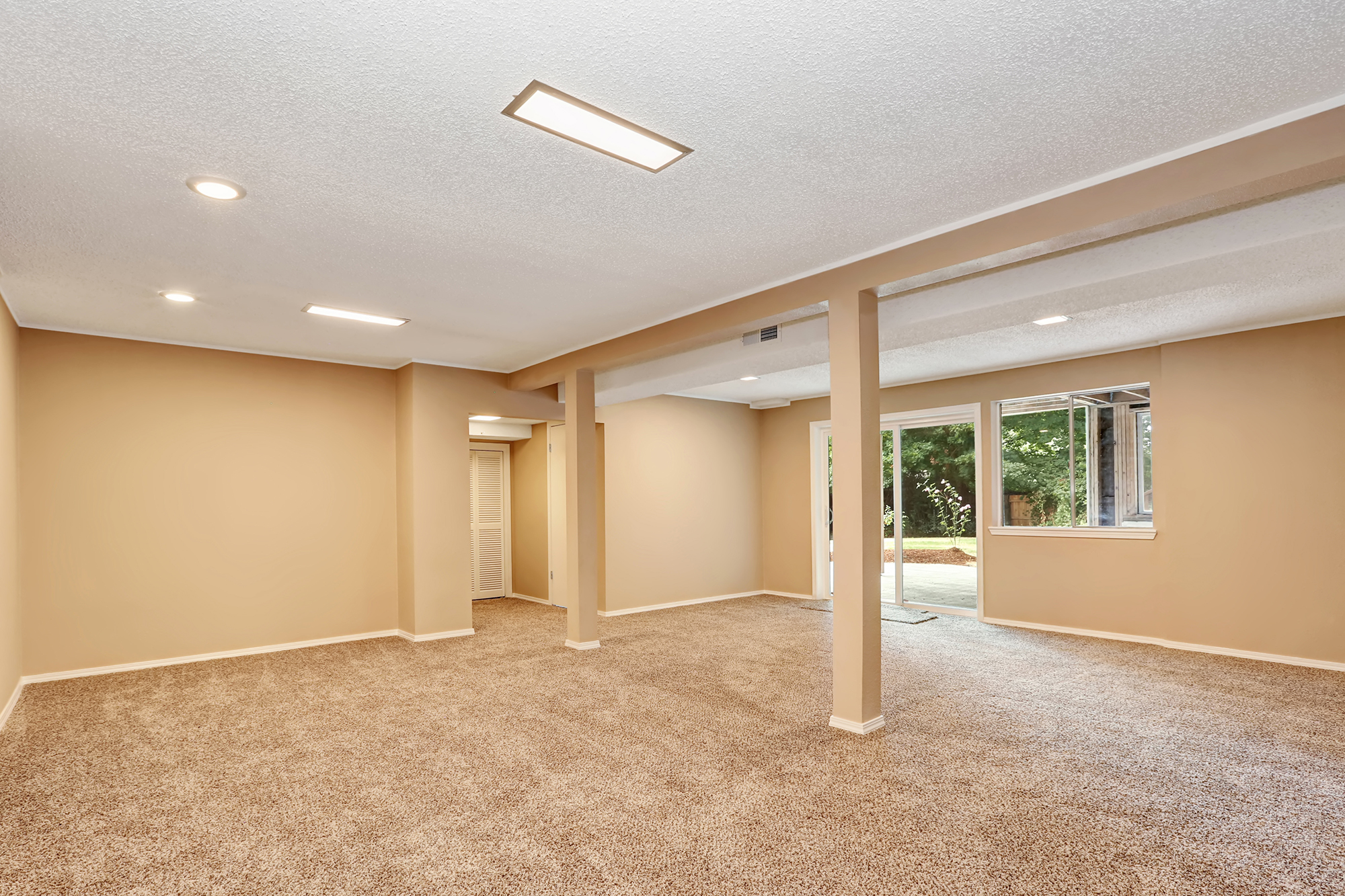 Spacious, carpeted room with beige walls, recessed lighting, and large windows overlooking a backyard.