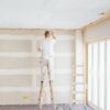 A person standing on a ladder paints the ceiling of a room under renovation. The unfinished drywall walls with visible joints highlight a project that shows when to hire a painter for professional results. Another empty ladder stands nearby next to large windows.