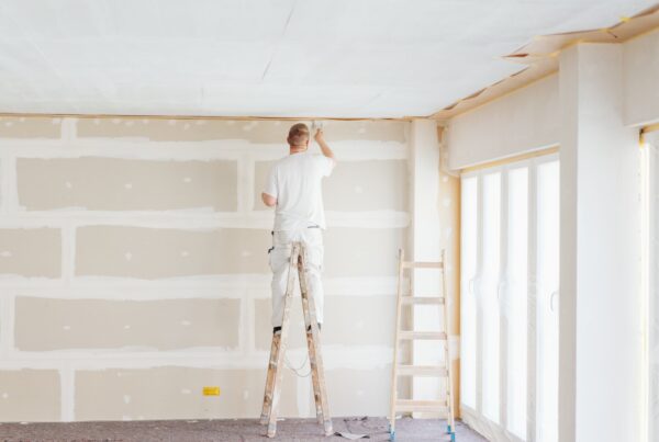 A person standing on a ladder paints the ceiling of a room under renovation. The unfinished drywall walls with visible joints highlight a project that shows when to hire a painter for professional results. Another empty ladder stands nearby next to large windows.
