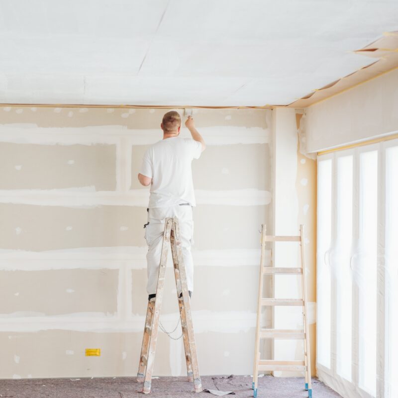 A person standing on a ladder paints the ceiling of a room under renovation. The unfinished drywall walls with visible joints highlight a project that shows when to hire a painter for professional results. Another empty ladder stands nearby next to large windows.