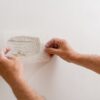 A person places a mesh patch over a hole in a white wall, showing how to patch drywall before painting and preparing the surface for repair.