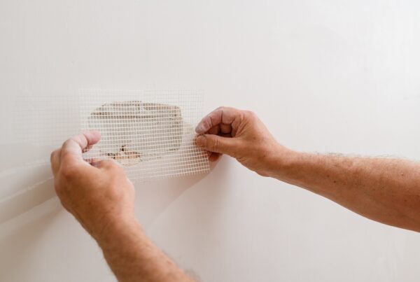 A person places a mesh patch over a hole in a white wall, showing how to patch drywall before painting and preparing the surface for repair.