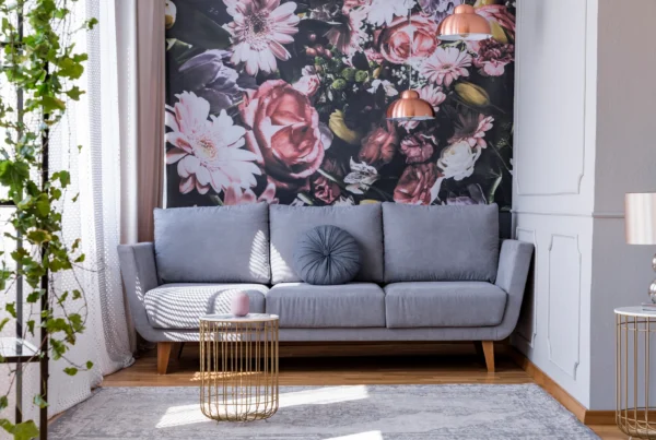 A modern living room with a gray sofa, round pillows, a gold wire-frame side table, a floral mural on the wall, and pink pendant lamps. Light enters through sheer curtains; greenery hangs on the left side of the image.