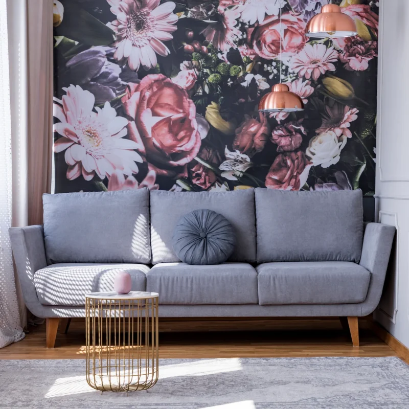 A modern living room with a gray sofa, round pillows, a gold wire-frame side table, a floral mural on the wall, and pink pendant lamps. Light enters through sheer curtains; greenery hangs on the left side of the image.