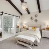 A modern bedroom with exposed dark ceiling beams, a bed with a beige headboard, two bedside tables with lamps, a bench at the foot of the bed, decorative mirrors on the wall, and a sliding door revealing a bathroom.