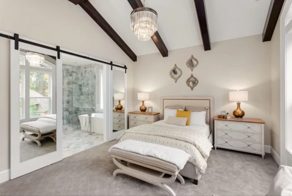 A modern bedroom with exposed dark ceiling beams, a bed with a beige headboard, two bedside tables with lamps, a bench at the foot of the bed, decorative mirrors on the wall, and a sliding door revealing a bathroom.