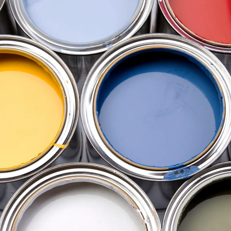 Open cans filled with vibrant Benjamin Moore paint colors by finish—yellow, blue, red, white, black, and other shades—are arranged closely together in a neat grid-like pattern.