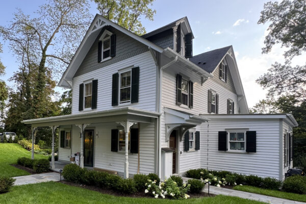 A two-story white house with black shutters, a covered front porch, and well-kept green lawn stands proudly, featured in our Residential Painting Portfolio, set against trees and a sunny, partly cloudy sky.