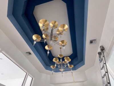 A modern ceiling from our Residential Painting Portfolio features a geometric blue recessed design with two silver and gold chandeliers. A ladder stands nearby, while large windows and archways brighten the room.