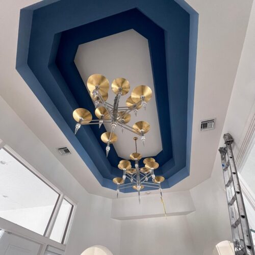 A modern ceiling from our Residential Painting Portfolio features a geometric blue recessed design with two silver and gold chandeliers. A ladder stands nearby, while large windows and archways brighten the room.