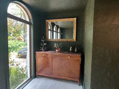 A small hallway with textured dark green walls showcases this Residential Painting Portfolio. A large arched window, a wooden sideboard beneath a gold-framed mirror, and decorative items complete the look atop light wood flooring.