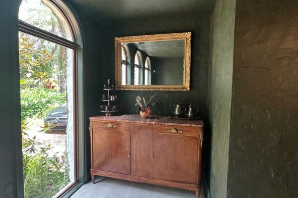 A small hallway with textured dark green walls showcases this Residential Painting Portfolio. A large arched window, a wooden sideboard beneath a gold-framed mirror, and decorative items complete the look atop light wood flooring.