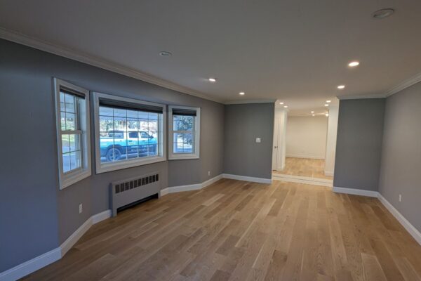 A spacious, empty room with light wood flooring, gray walls, recessed ceiling lights, a large bay window, and an open doorway leading to another room—perfect for showcasing your Residential Painting Portfolio. A parked vehicle is visible outside the window.