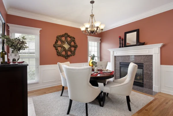 A dining room with four white upholstered chairs around a round table, set for a meal. The terracotta walls, carefully achieved by painting over dark colors, feature white trim, a decorative mirror, and a cozy fireplace with candles.