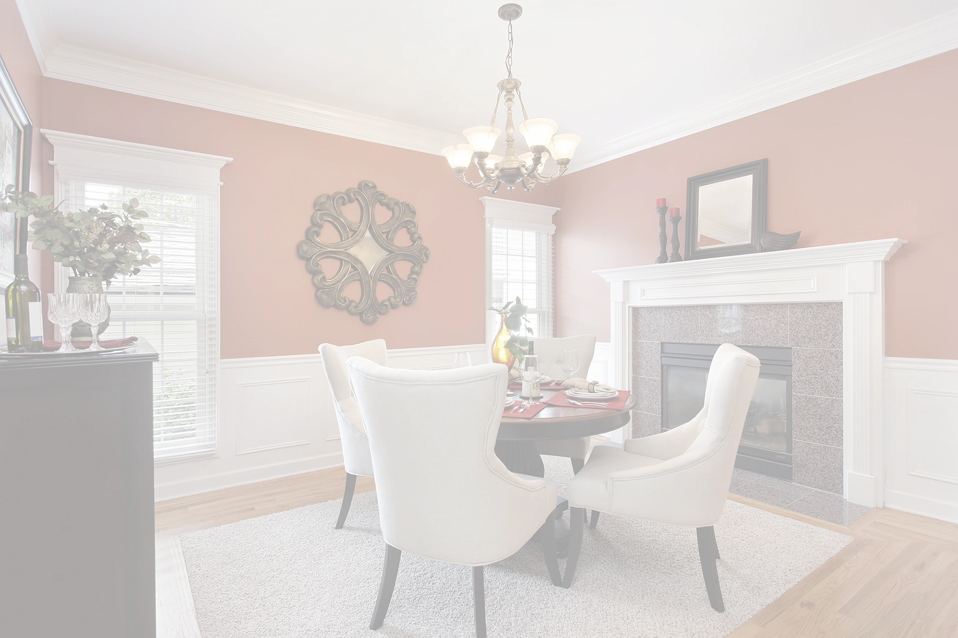 A dining room with a round table set for four, surrounded by white upholstered chairs, a fireplace with a mantle, wall mirror, chandelier, and large windows letting in natural light—perfect for painting over dark colors to refresh the space.