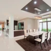 Modern dining room with a long table set for eight, white chairs, brown rug, pendant lights, large windows with curtains, and an open view into the kitchen and living area. The space is bright and spacious.