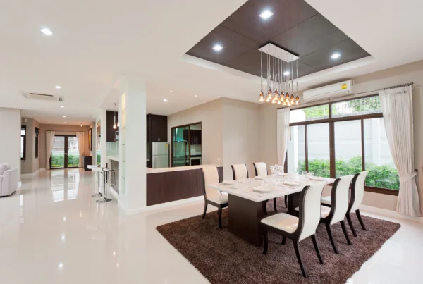 Modern dining room with a long table set for eight, white chairs, brown rug, pendant lights, large windows with curtains, and an open view into the kitchen and living area. The space is bright and spacious.