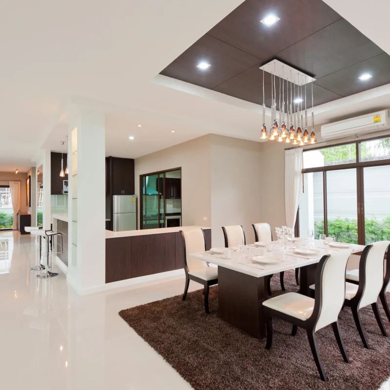Modern dining room with a long table set for eight, white chairs, brown rug, pendant lights, large windows with curtains, and an open view into the kitchen and living area. The space is bright and spacious.